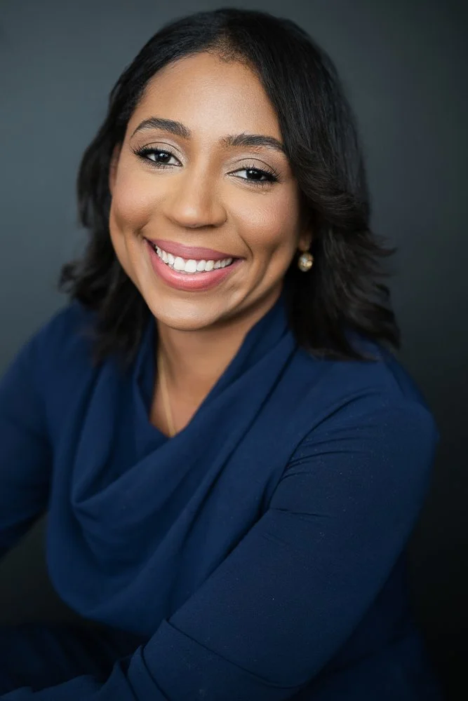 A smiling woman with dark, shoulder-length hair and pearl earrings, wearing a dark blue top against a dark background.