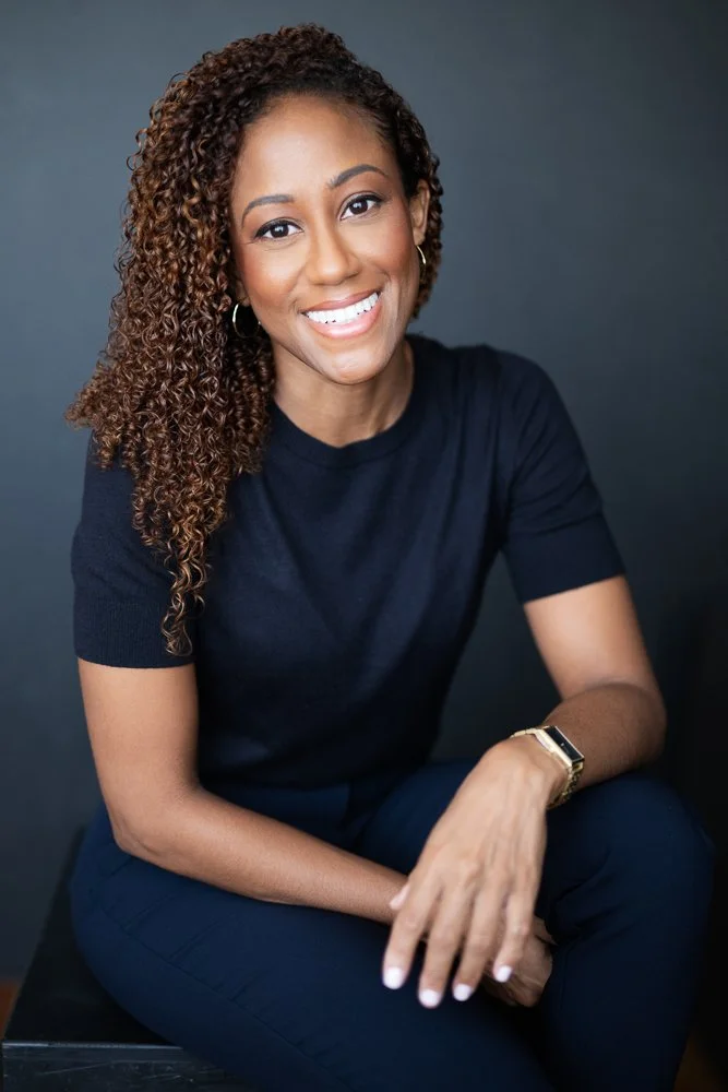 Smiling woman with curly hair, wearing a black t-shirt, sitting against a dark gray background.