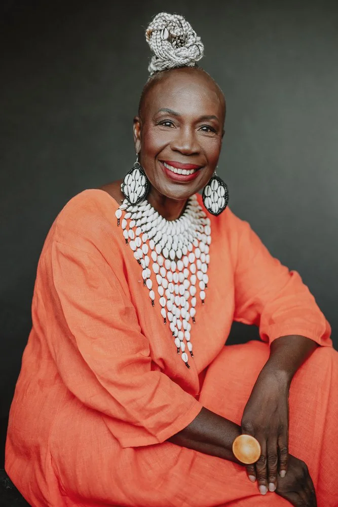 A smiling woman dressed in an orange outfit with large white jewelry, including a layered necklace, earrings, and a headwrap, sitting against a dark background.