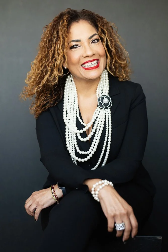 Woman with curly hair smiling, wearing a black blazer and multiple strands of pearl necklaces, with a pearl bracelet, ring, and printed black and white flower brooch, against gray background.