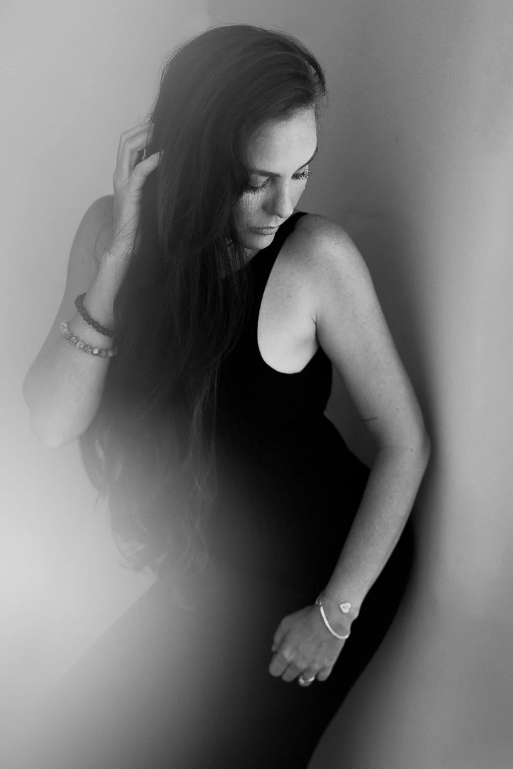 Black and white portrait of a woman with long hair, wearing a sleeveless dress, looking down gently, standing against a plain wall.