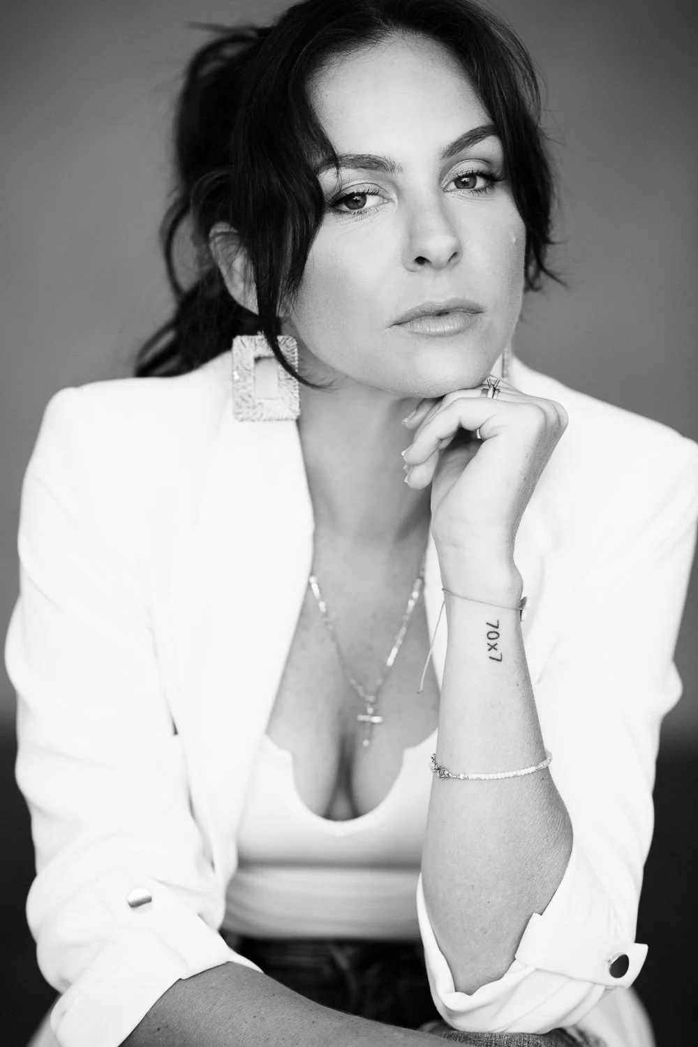 A black and white portrait of a woman with dark, wavy hair, wearing a white blazer and large earrings, gazing at the camera with her hand resting on her chin.