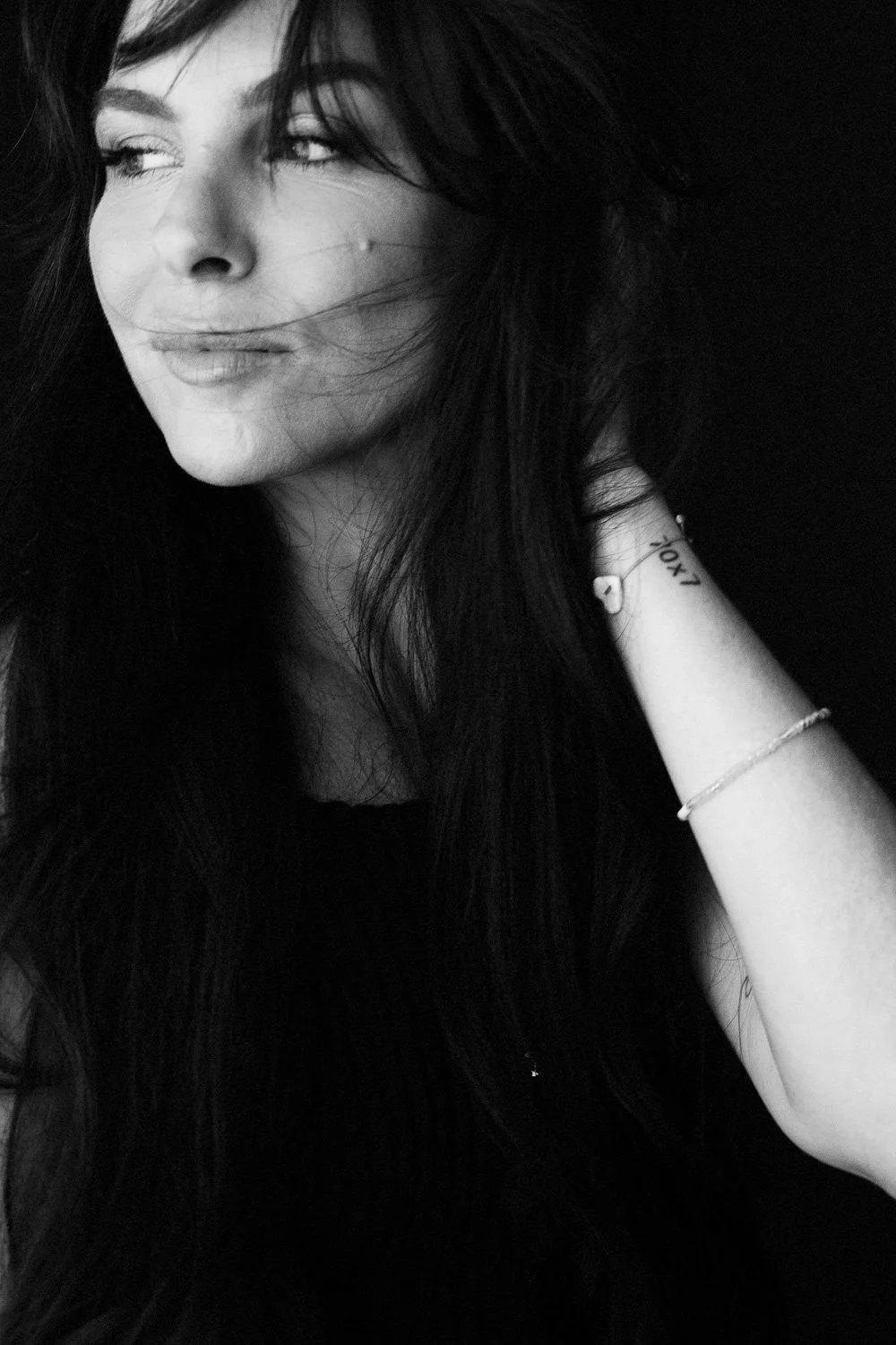 A black-and-white portrait of a woman with long dark hair, looking to her left with a slight smile, wearing bracelets and a ring, with her hand touching her neck.