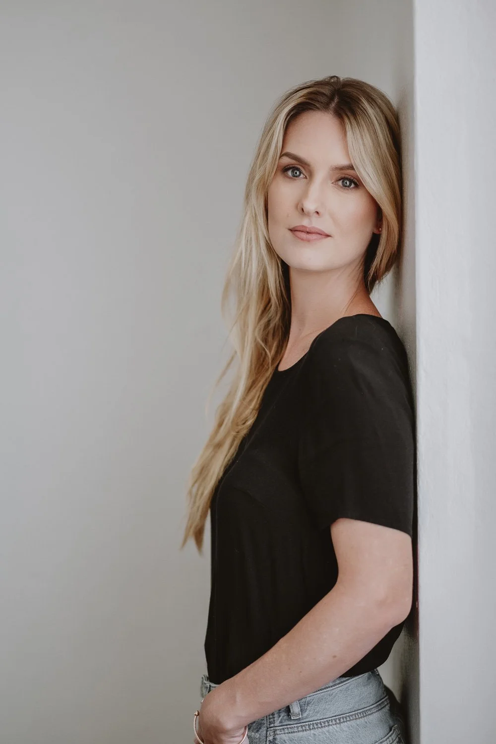 A woman with long blonde hair, wearing a black top, standing against a white wall, looking directly at the camera.