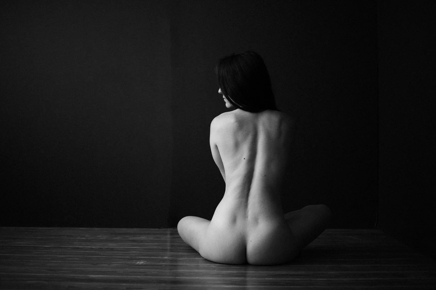 A black and white photo of a naked woman sitting on the wooden floor with her back facing the camera, against a dark plain background.