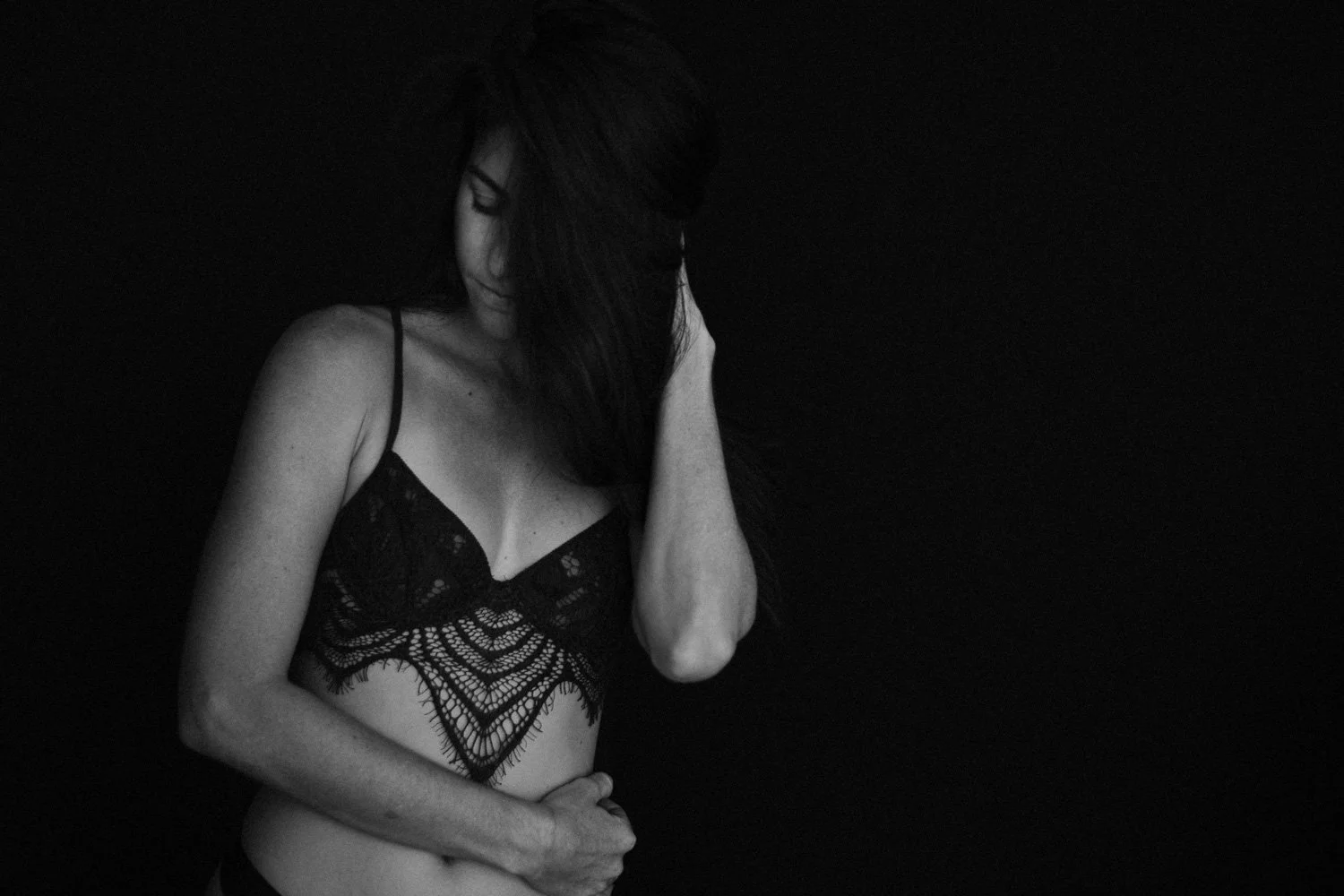 A woman with long dark hair wearing a lace black top, standing against a dark background, with her eyes closed and her hand touching her face.