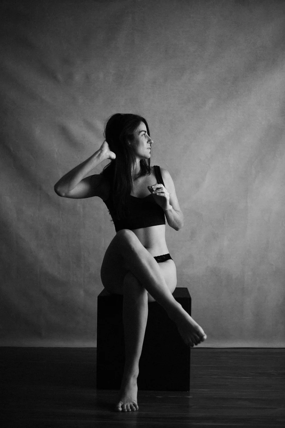 A black-and-white photo of a woman in black lingerie sitting on a black box, with one leg crossed over the other, looking to the right, with one hand behind her head and the other holding a small object.