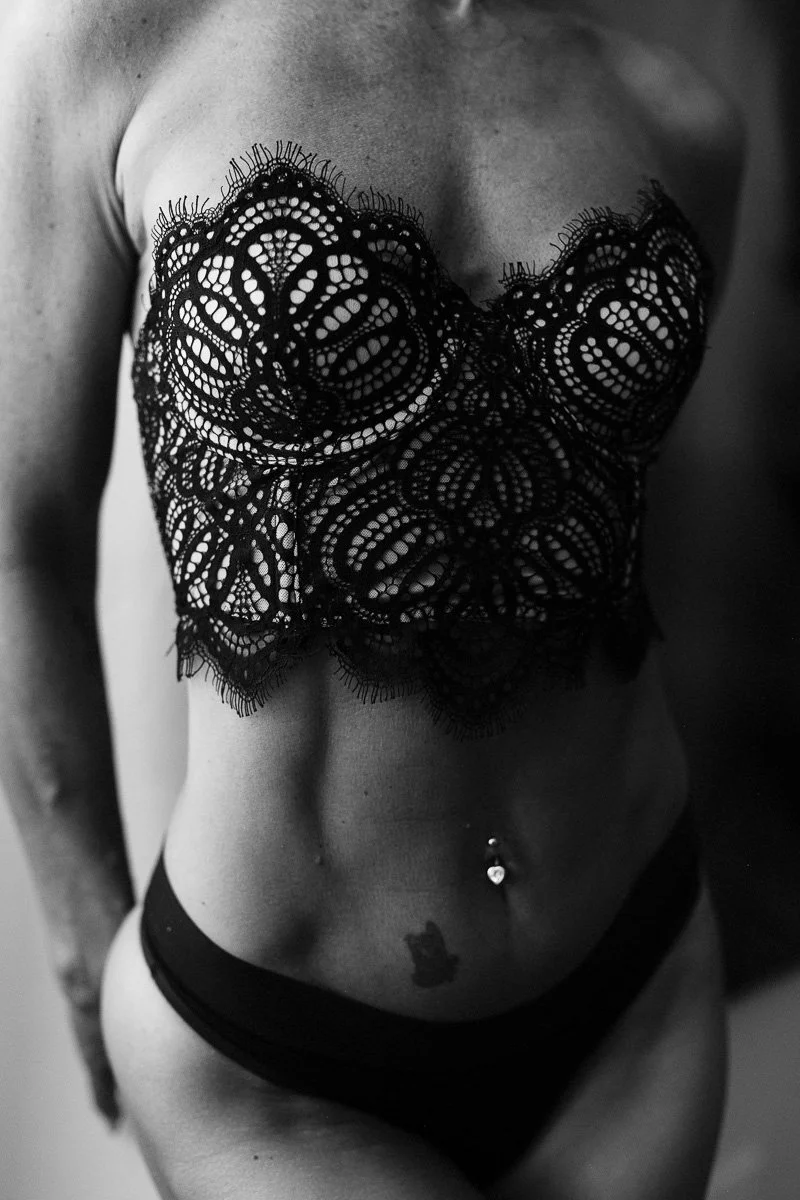 A woman wearing lace lingerie, showing a navel piercing and a tattoo on her lower abdomen.