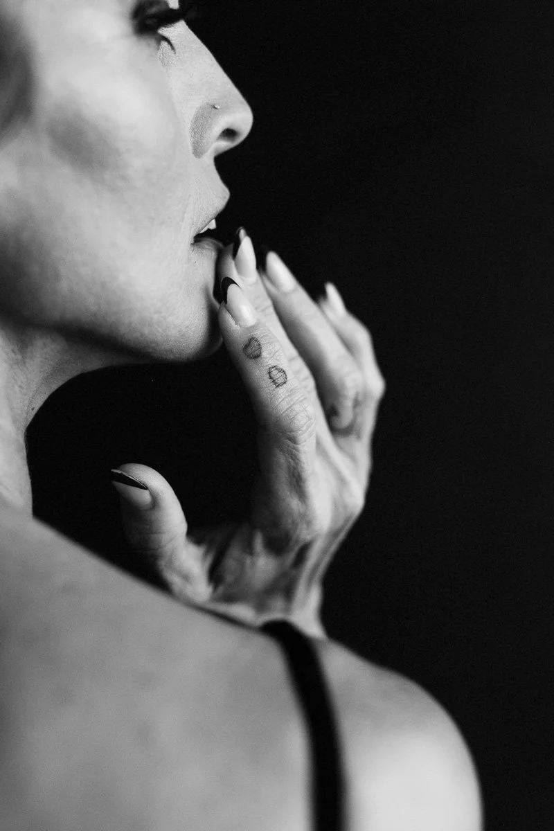 Close-up black and white photo of a woman with her eyes closed, touching her lips with her fingers, which have tattoos and dark nail polish.