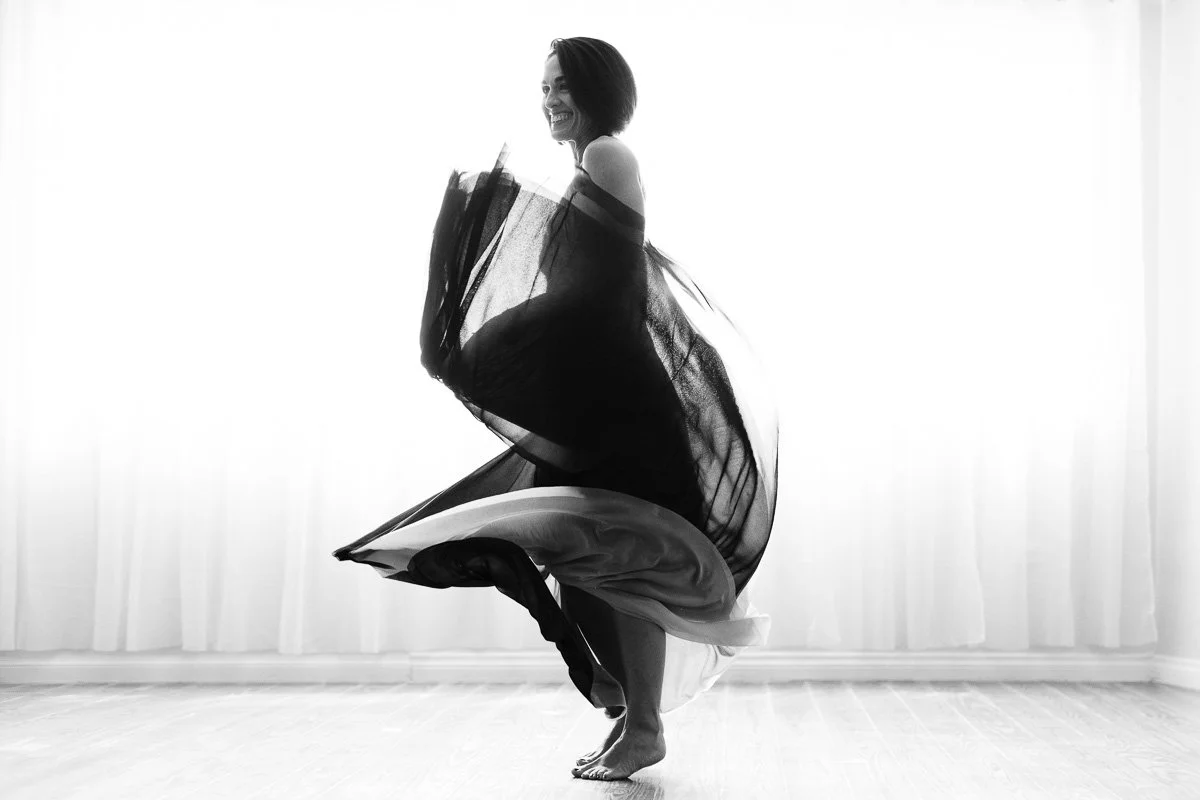 A woman smiling and dancing in a black and white dress in front of a bright window.