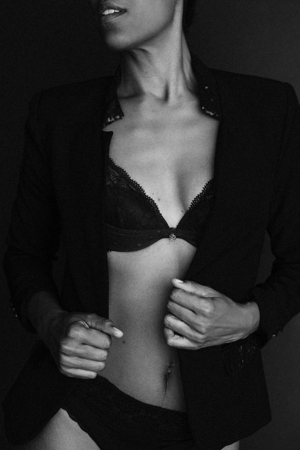 A woman in a black blazer, lace bra, and underwear with a navel piercing, posing against a dark background.