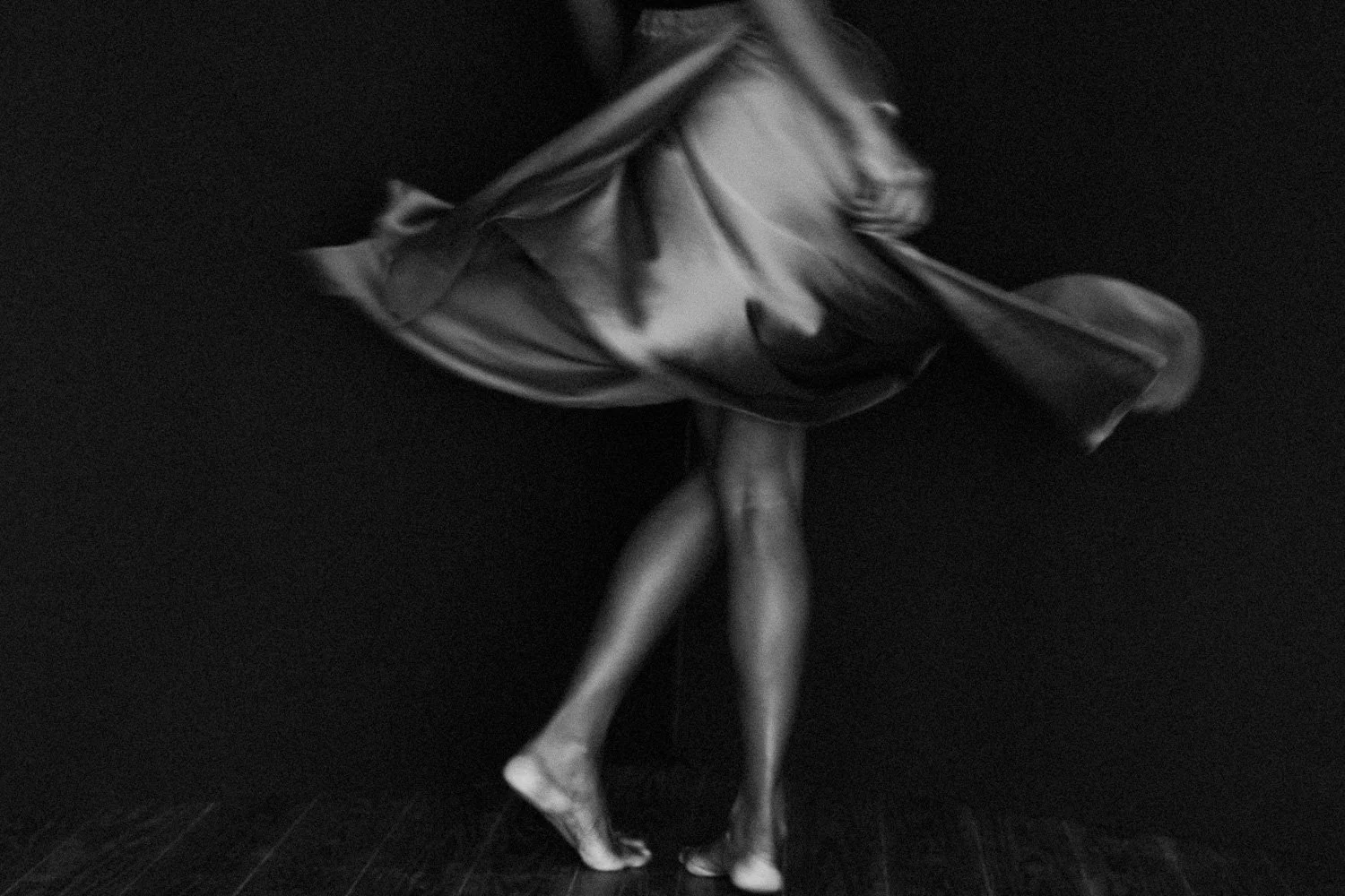 Black and white photo of a person with long hair, standing on a dark floor, with their legs crossed at the ankles, facing away from the camera, and wearing a flowing dress.
