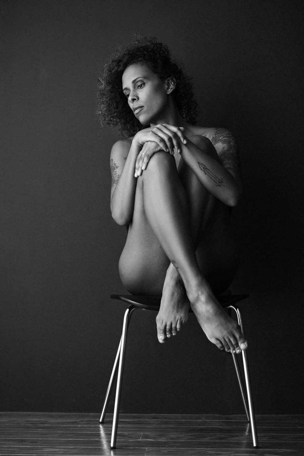 A black and white photo of a nude woman with tattoos, sitting cross-legged on a chair against a dark background, with her arms wrapped around her knees and her head bowed slightly.