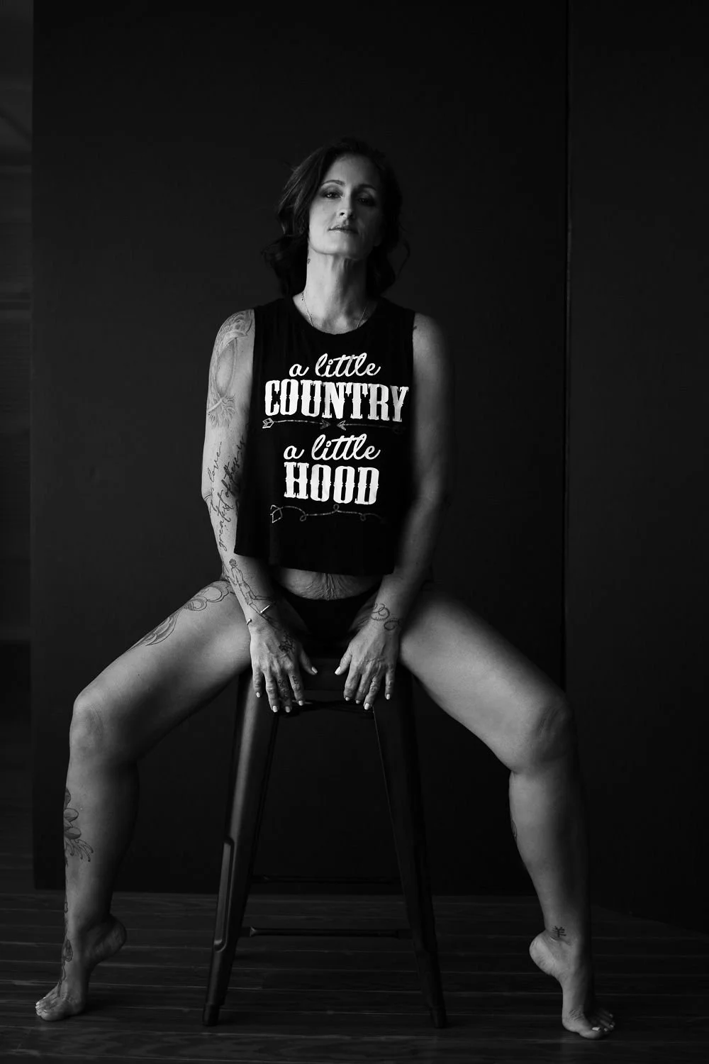 A woman with tattoos sitting on a stool with legs apart, against a dark background, wearing a sleeveless shirt and underwear, in black and white photograph.