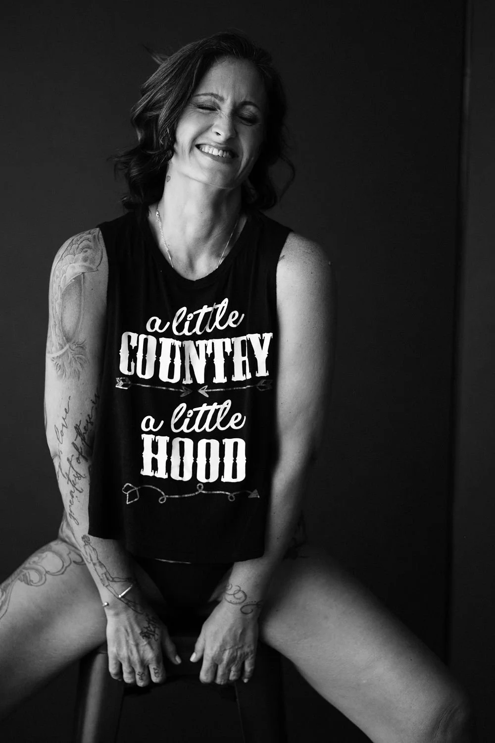 A smiling woman with tattoos wearing a sleeveless shirt that says 'a little COUNTY, a little HOOD' in black and white photo.
