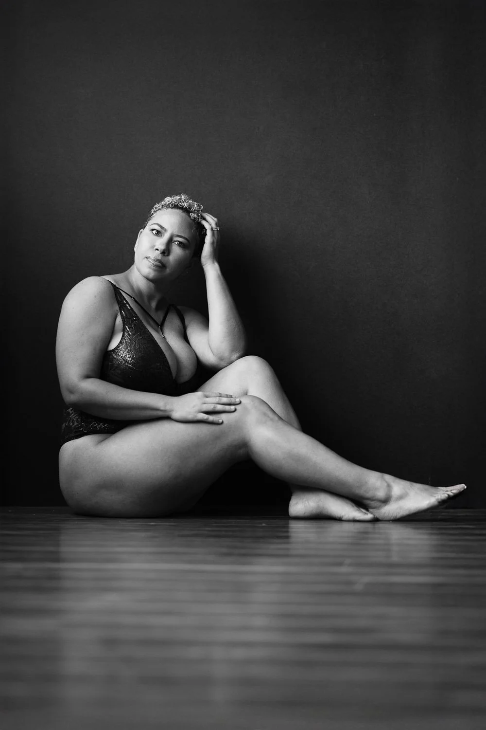 Black and white photo of a woman sitting on the floor with her legs crossed, wearing lingerie, against a plain dark wall, with her hand resting on her head and looking thoughtfully into the distance.