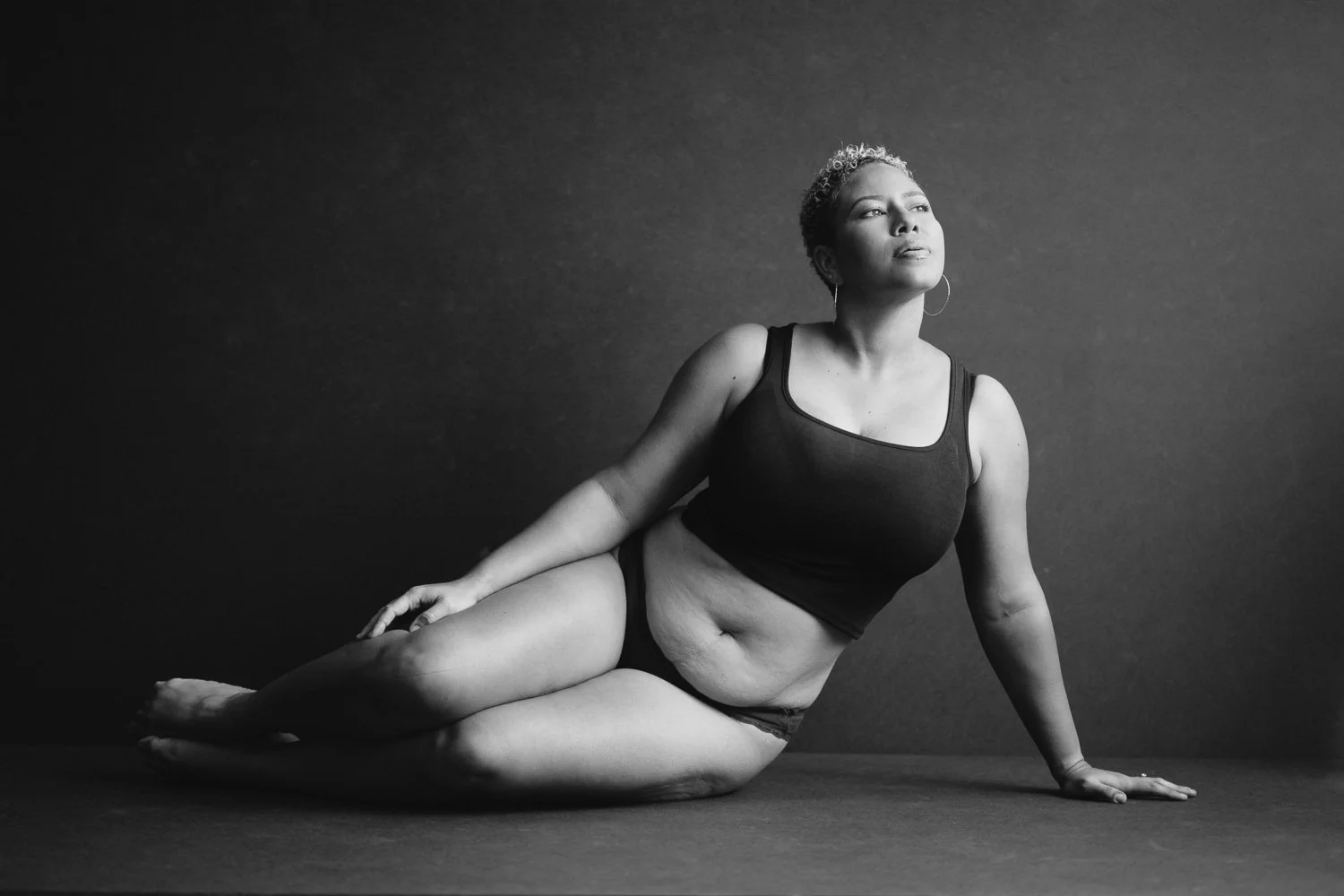 Black and white photo of a woman sitting on the floor in a yoga pose, with her torso upright, one arm supporting her, and her face turned slightly upward, against a plain dark background.