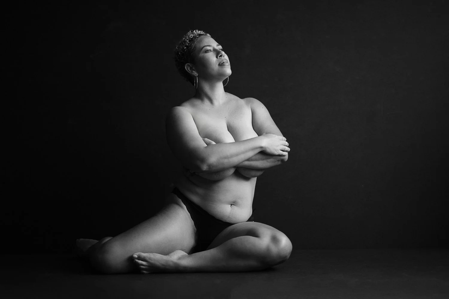 Black and white photo of a woman kneeling on the floor, topless, crossing her arms over her chest, with a contemplative expression, against a dark background.