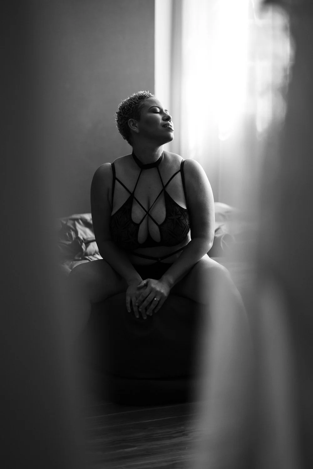 A woman with short hair sitting on a bench in front of a window, wearing lingerie with intricate strap details, lighting coming from the right side, black and white photo.