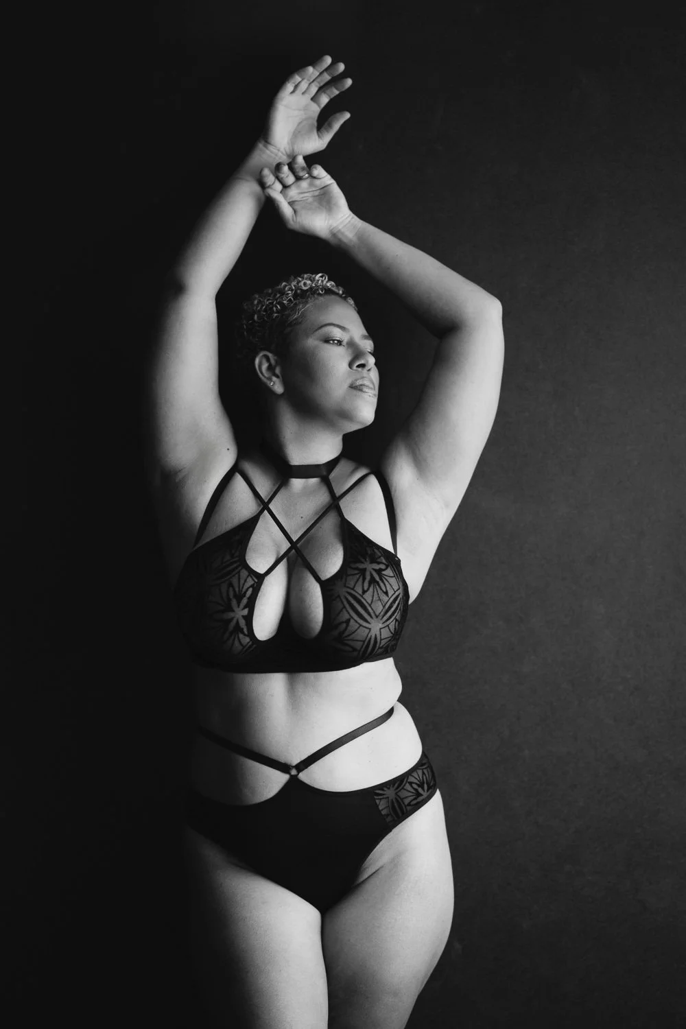 A woman in black lingerie with intricate cutout designs and straps, posing with arms raised and her eyes closed against a dark background.