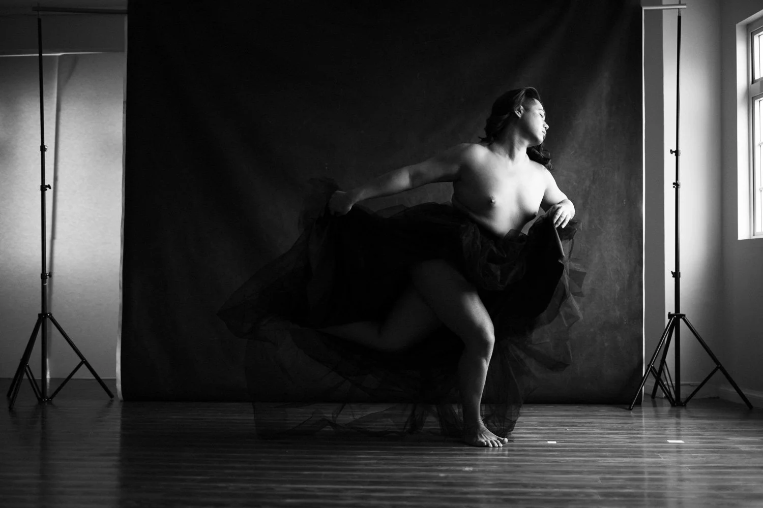 A woman dancing topless in a photography studio, wearing a black tulle skirt, against a dark backdrop, with studio lights and windows visible.