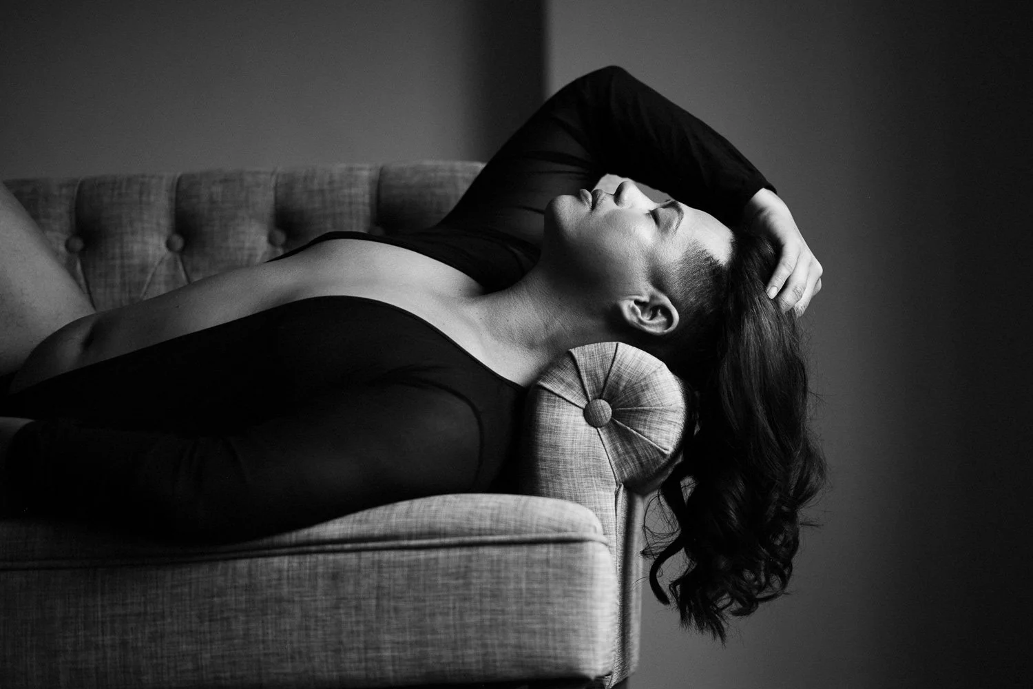 Black and white photo of woman lying on a sofa, with her head tilted back and eyes closed, hand resting on her forehead, wearing a long-sleeve top.