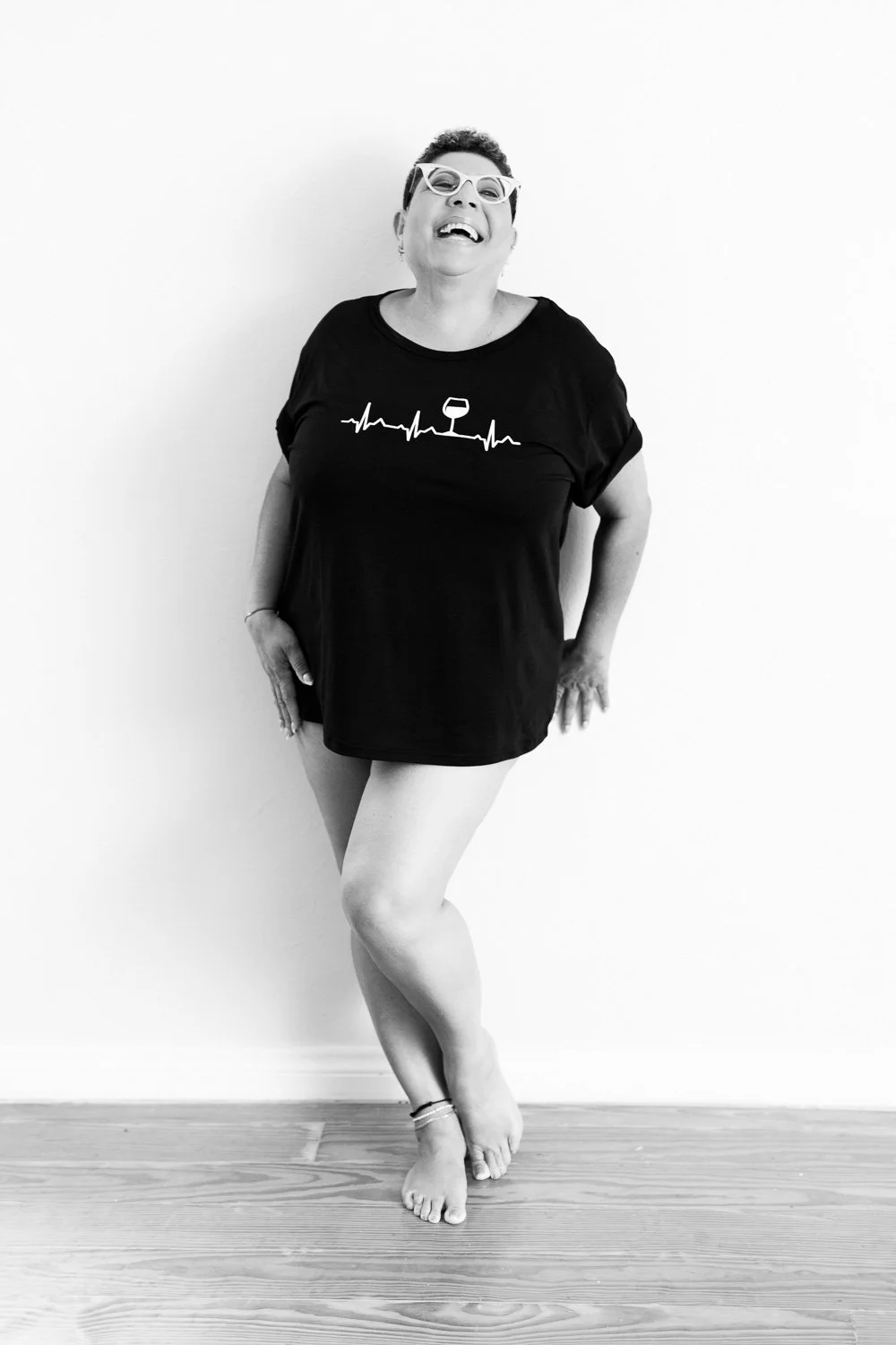 A woman with short hair, sunglasses, and a big smile, standing against a white wall, wearing a black t-shirt with a musical note design and shorts, with one foot crossed over the other.