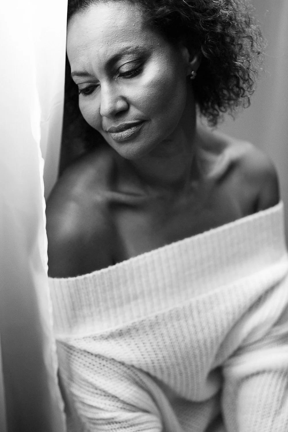 A black and white portrait of a woman with curly hair, wearing an off-the-shoulder sweater, with her eyes closed and a peaceful expression.