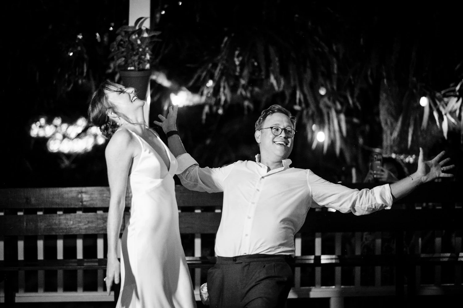 A black and white photo of a joyful couple dancing outdoors at night, with one person smiling broadly and the other laughing with head tilted back, surrounded by trees and string lights.