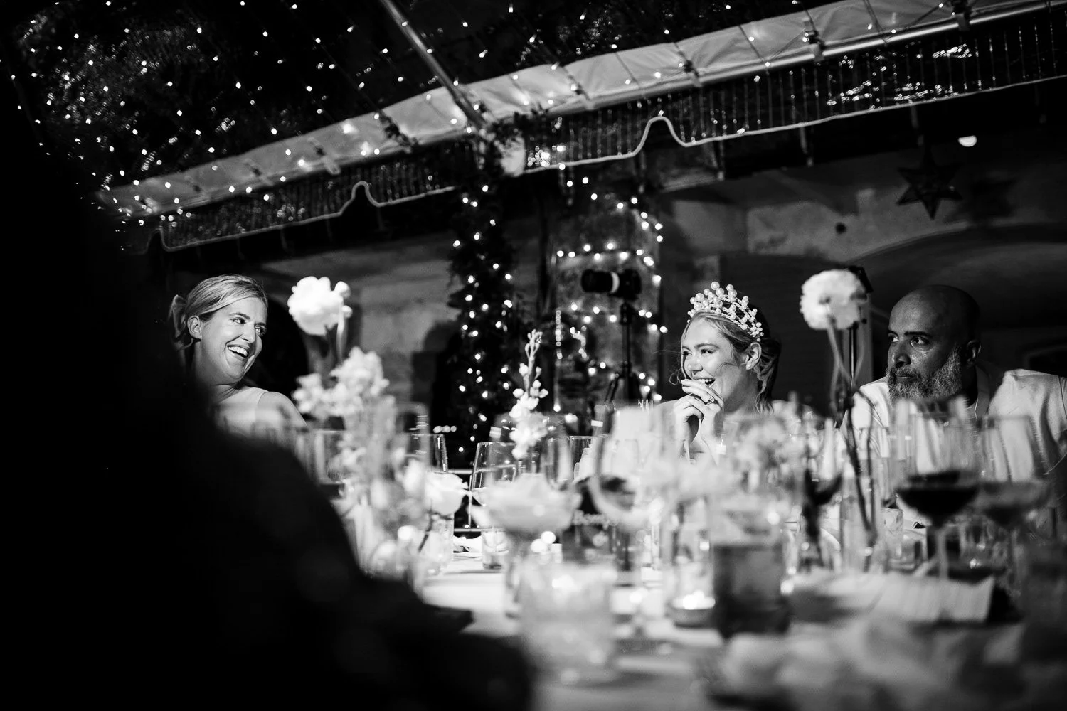 A black and white photo of a joyful woman wearing a crown, sitting at a decorated table during a celebration. She is smiling and laughing with two other people visible nearby. The setting includes string lights, floral centerpieces, and a festive atm