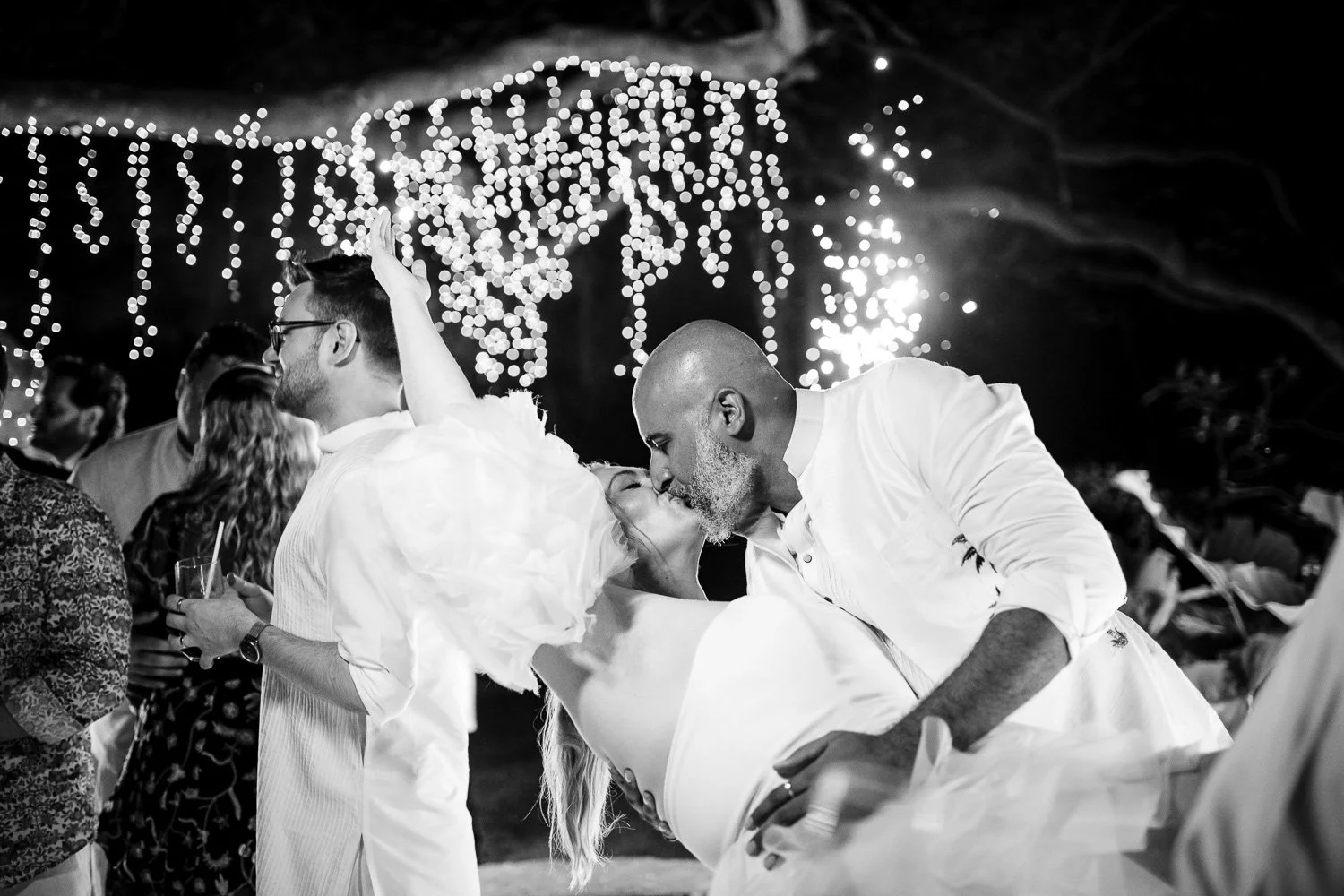 People dancing and kissing at a festive outdoor event with string lights in the background