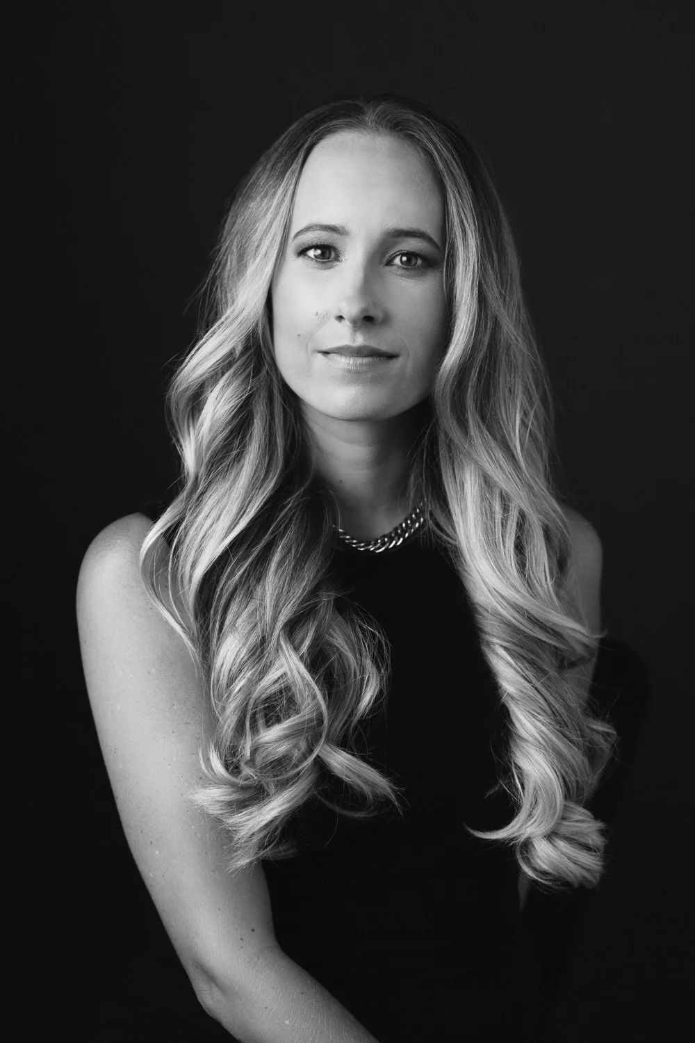 Black and white portrait of a woman with long, wavy hair wearing a sleeveless top and a chain necklace, looking directly at the camera.