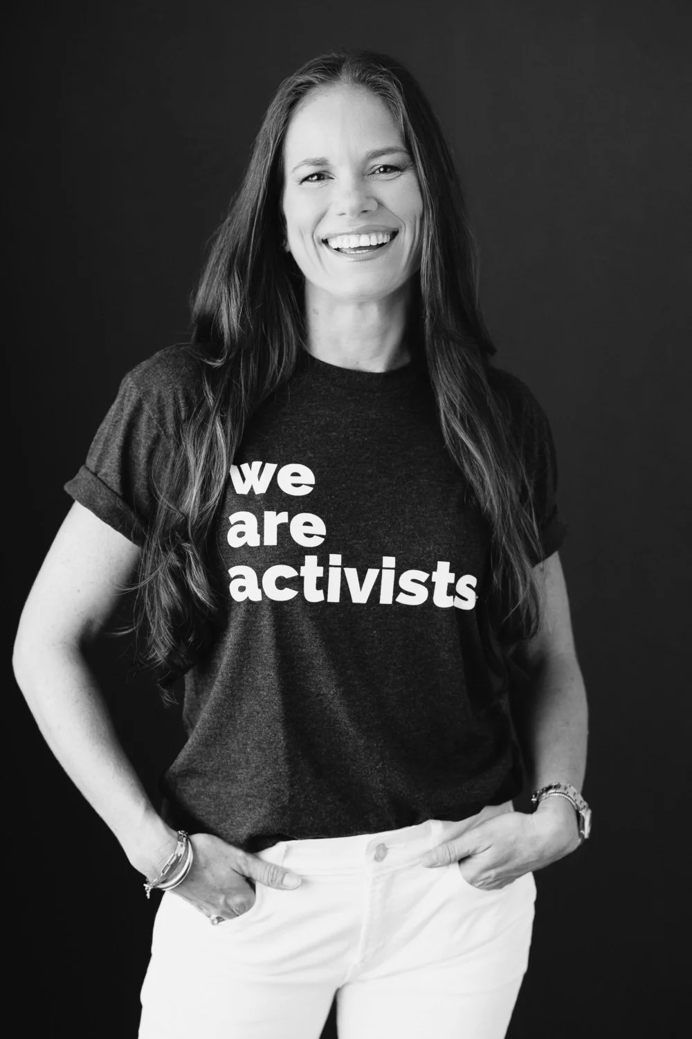 A woman smiling, wearing a dark t-shirt that says "we are activists", against a dark background.
