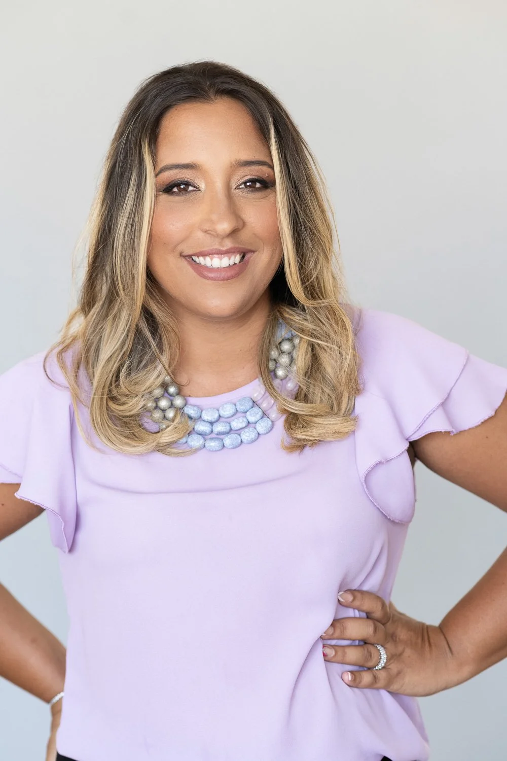 A woman with long, wavy blonde hair, wearing a light purple top with ruffled sleeves, a pearl necklace, and a ring, posing with her hand on her hip and smiling at the camera.
