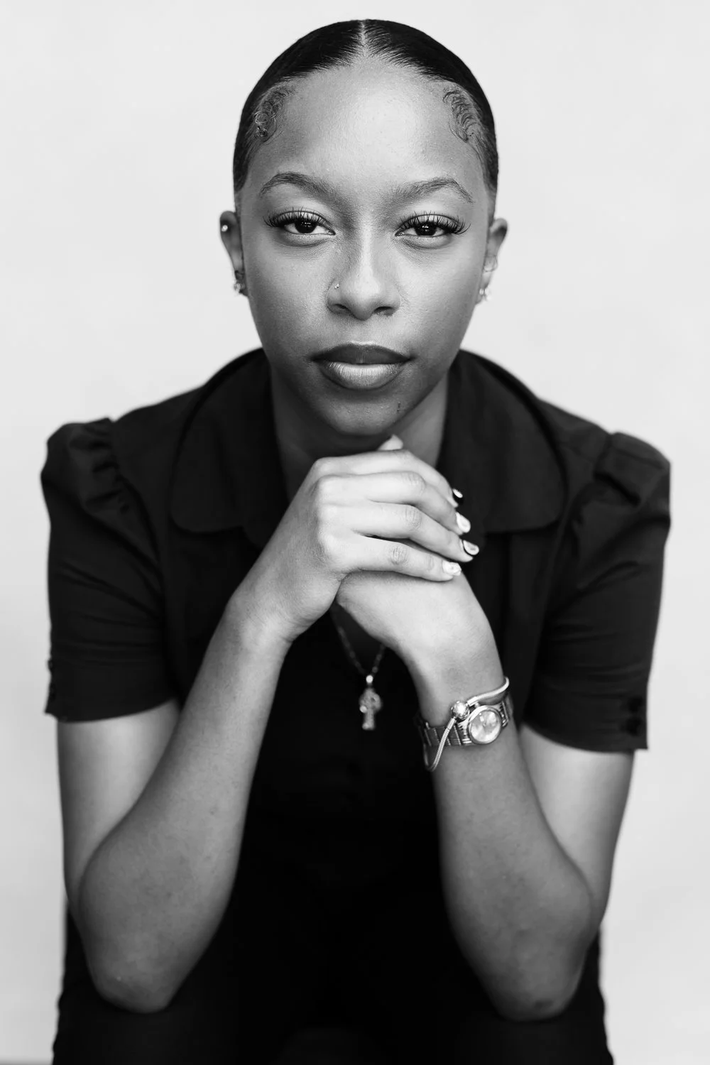 Black and white portrait of a woman with her hands clasped under her chin, wearing a watch and jewelry, looking directly at the camera with a neutral expression.