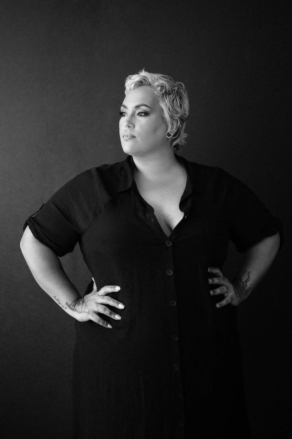 A woman with short, curly blonde hair standing with hands on hips against a dark background.