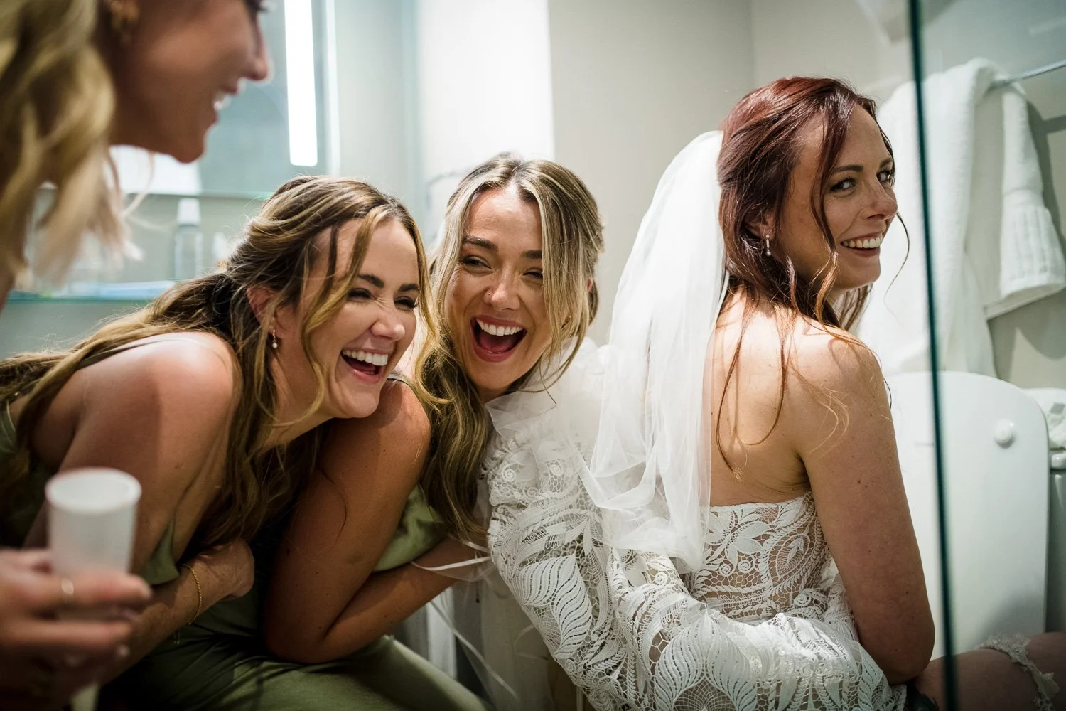 Four women, one wearing a wedding dress, are smiling and laughing together in a bathroom