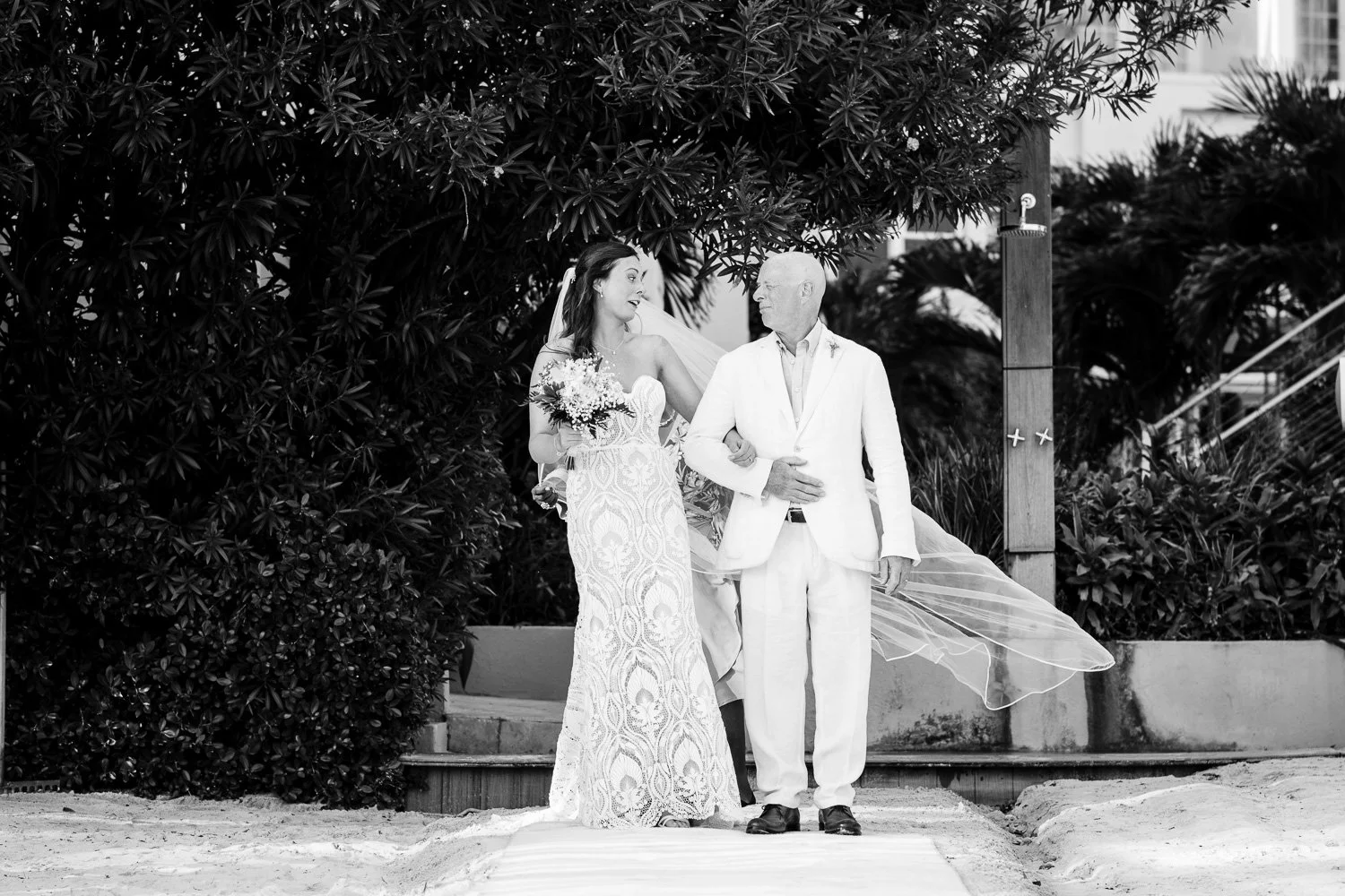 A bride holding a bouquet walking arm-in-arm with an older man, likely her father, on a beach during a wedding ceremony. The bride is wearing a lace wedding dress and a veil, and the man is dressed in a white suit. They are under a large tree with fo