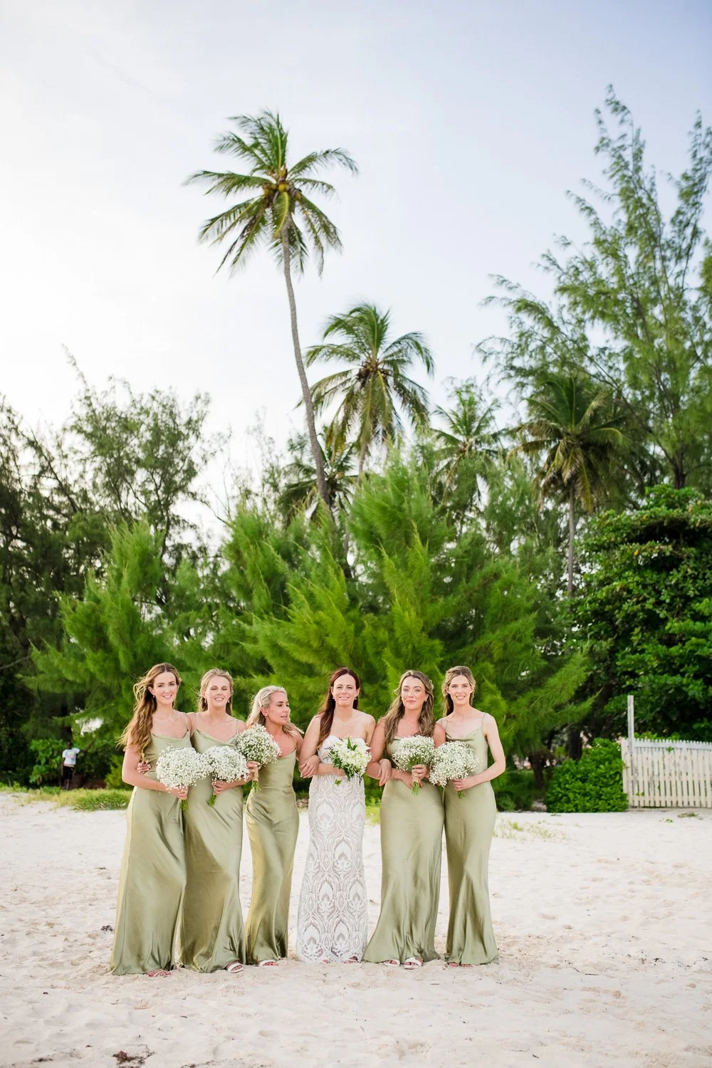 Group of six women in bridesmaid dresses and wedding dress holding bouquets on a beach with palm trees in the background.