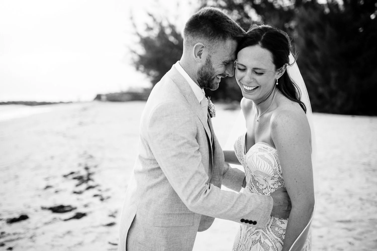 Black and white photo of a smiling wedding couple on the beach, with the groom in a light-colored suit and the bride in a lace dress, leaning their foreheads together.