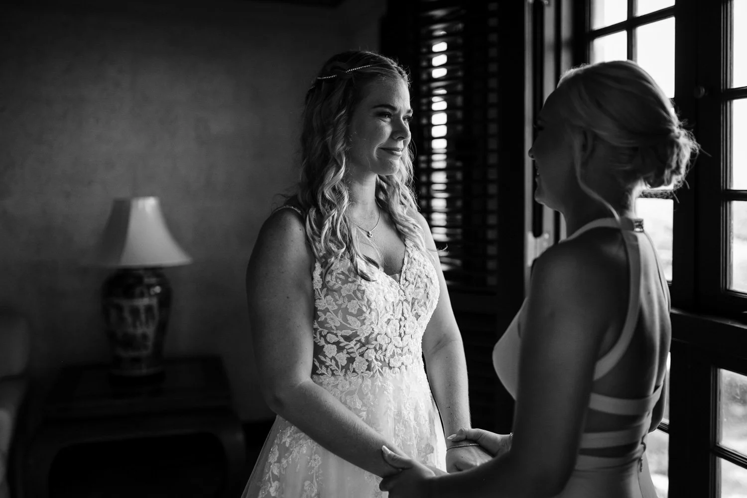 A black and white photo of two women backstage before a wedding, holding hands, with one in a wedding dress and the other in a sleeveless dress, smiling at each other near a window.