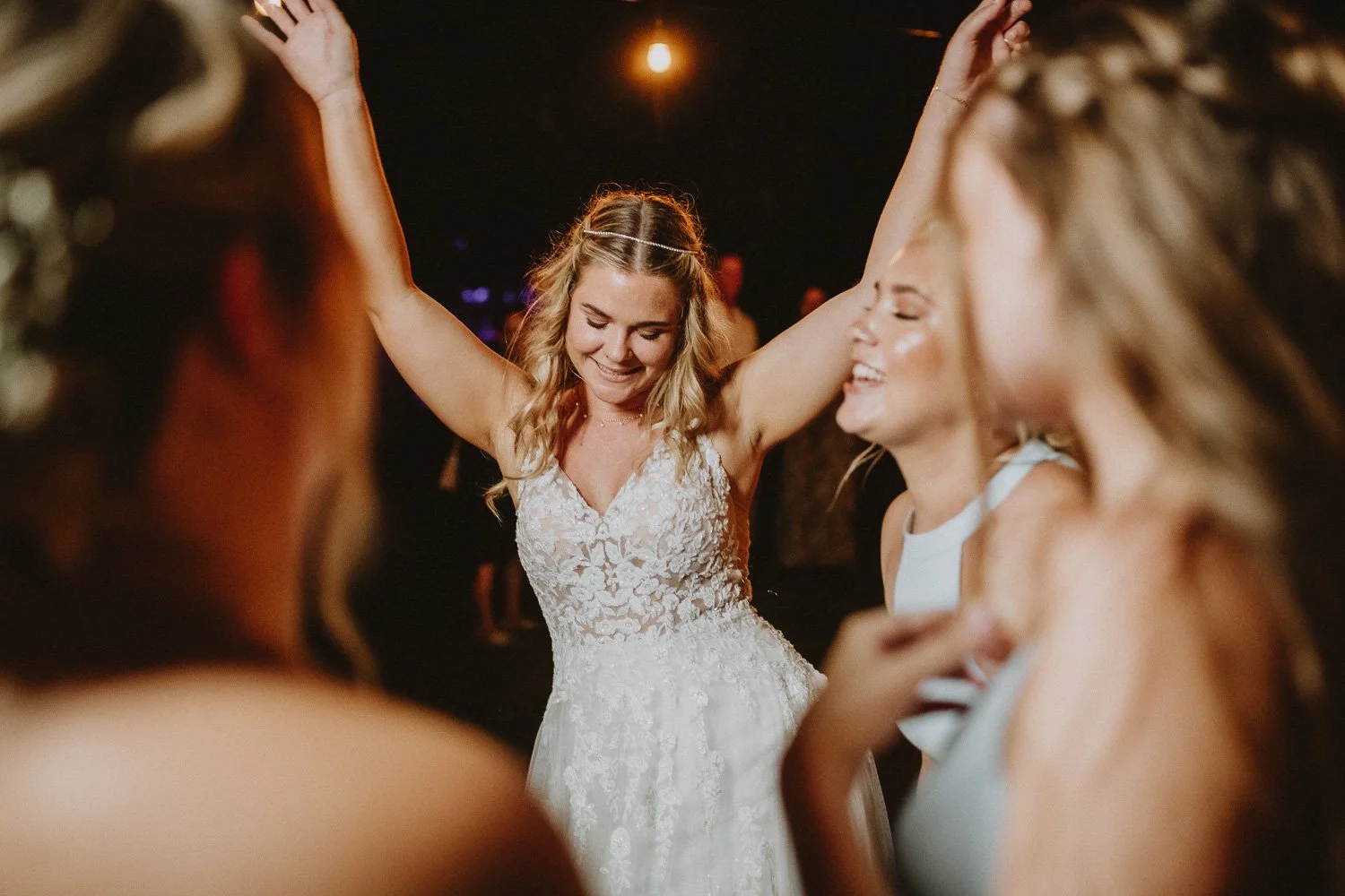 A woman in a wedding dress dancing with her arms raised at a celebration, surrounded by friends.