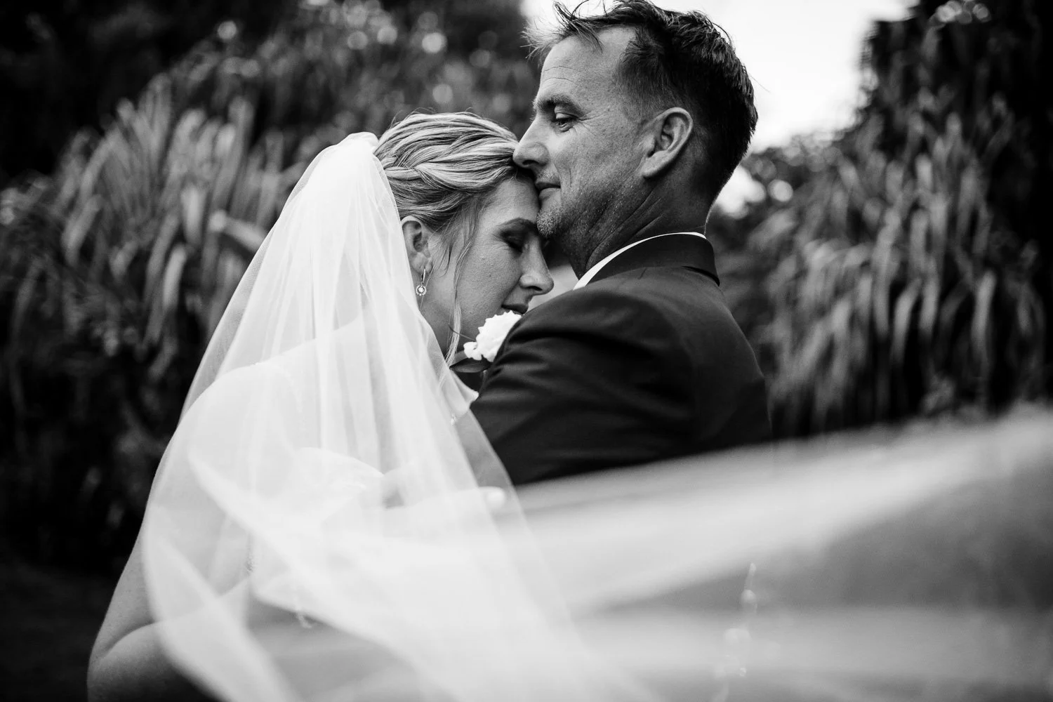 Black and white photo of a woman in a wedding dress with a veil and pearl earrings, leaning her head on a man in a tuxedo, both with eyes closed, in a tender embrace outdoors.