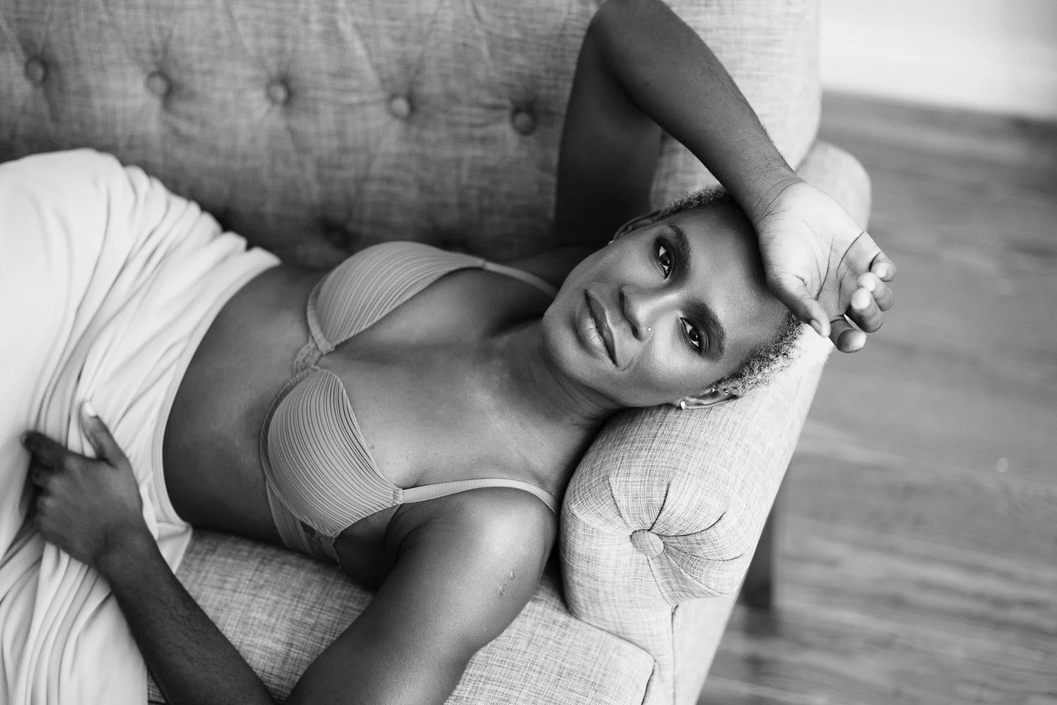 A woman with short curly hair reclining on a sofa, wearing a striped bra and light-colored skirt, resting her right arm on her forehead and looking at the camera. The setting is indoors with wooden flooring.