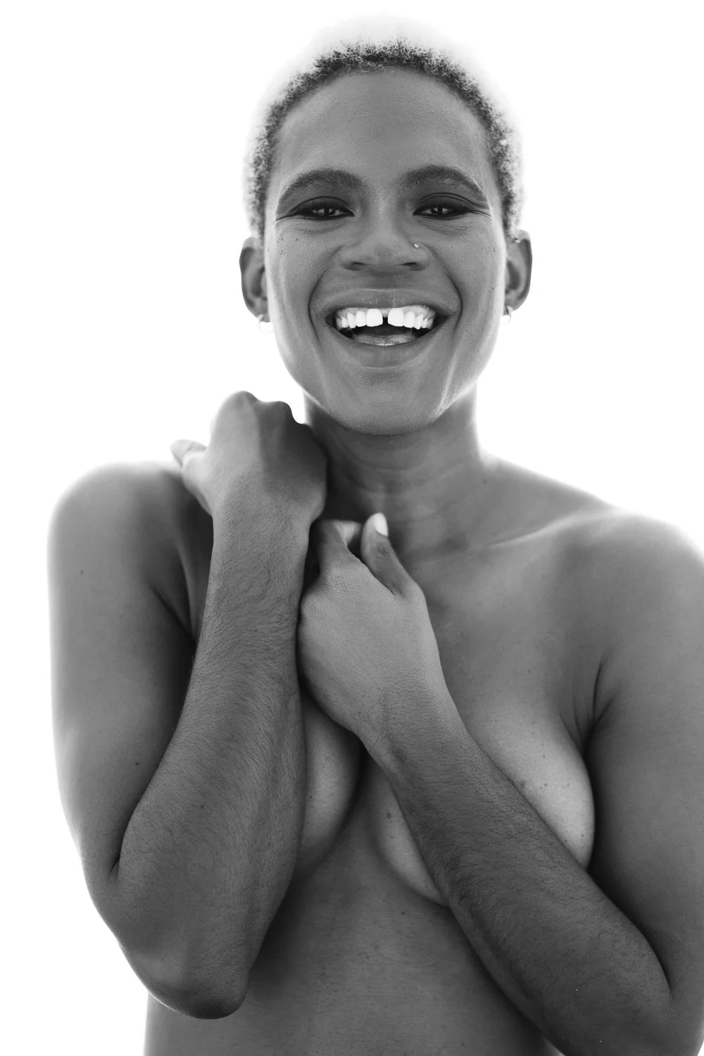 Black and white portrait of a smiling woman with short hair, no shirt, pulling her shoulder with one hand, and clasping her hands over her chest, against a bright background.