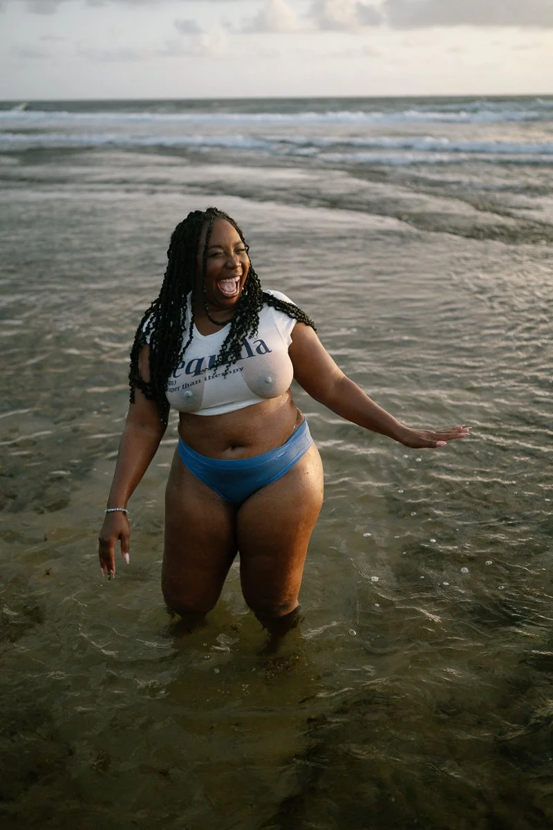 A woman standing in the ocean water, smiling and enjoying her time at the beach, with waves and cloudy sky in the background.