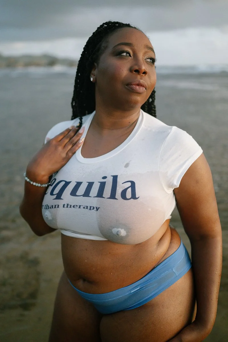 A woman at the beach wearing a white T-shirt that says 'tequila' and blue underwear, with one hand on her chest, gazing thoughtfully into the distance.