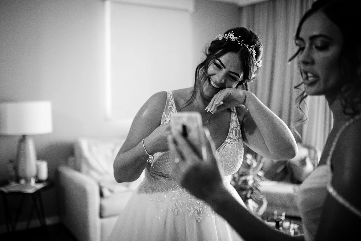 A bride in a wedding dress laughing and looking at her phone, standing next to another woman in a hotel room with curtains and a sofa.