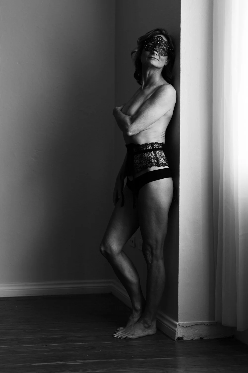 Black and white photo of a topless woman wearing lace underwear and a decorative mask, standing in a corner, facing slightly to the side with arms crossed.