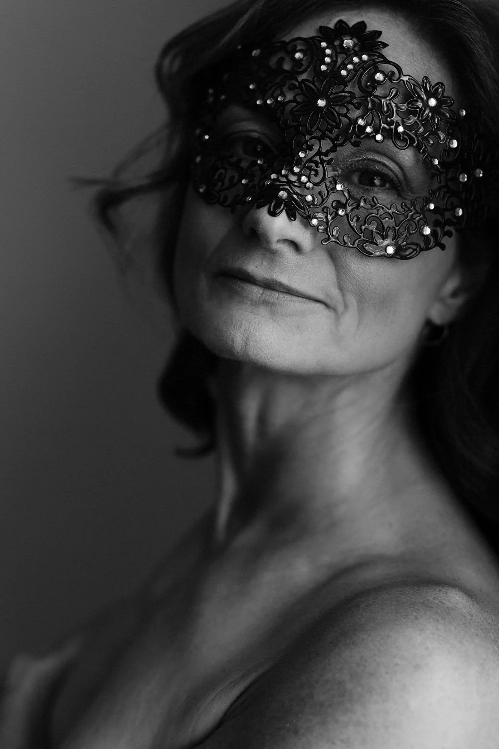 A woman with dark hair wearing an ornate black lace mask decorated with pearls, smiling gently, in black and white.